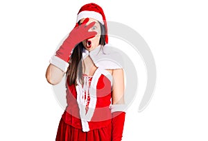 Young beautiful caucasian woman wearing santa claus costume peeking in shock covering face and eyes with hand, looking through