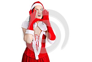 Young beautiful caucasian woman wearing santa claus costume shouting angry out loud with hands over mouth