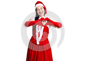 Young beautiful caucasian woman wearing santa claus costume smiling in love doing heart symbol shape with hands