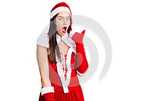 Young beautiful caucasian woman wearing santa claus costume surprised pointing with hand finger to the side, open mouth amazed