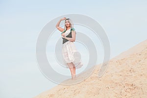 Young beautiful Caucasian woman in white dress and brown leather waistcoat posing in desert landscape with sand.