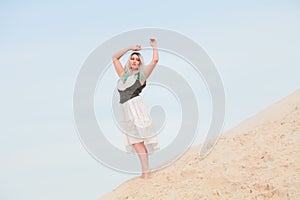 Young beautiful Caucasian woman in white dress and brown leather waistcoat posing in desert landscape with sand.