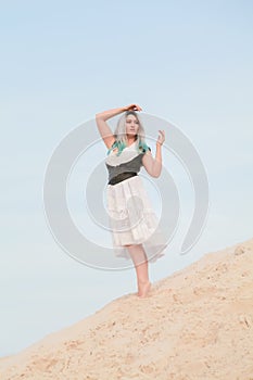 Young beautiful Caucasian woman in white dress and brown leather waistcoat posing in desert landscape with sand.