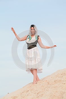 Young beautiful Caucasian woman in white dress and brown leather waistcoat posing in desert landscape with sand.