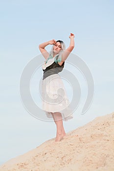 Young beautiful Caucasian woman in white dress and brown leather waistcoat posing in desert landscape with sand.