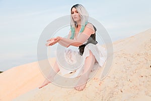Young beautiful Caucasian woman in white dress and brown leather waistcoat posing in desert landscape with sand.