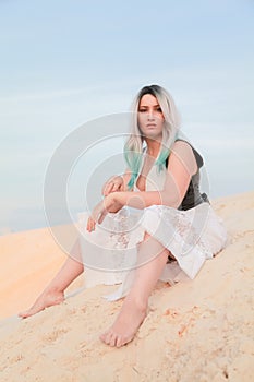 Young beautiful Caucasian woman in white dress and brown leather waistcoat posing in desert landscape with sand.