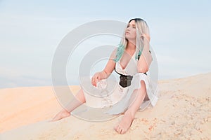 Young beautiful Caucasian woman in white dress and brown leather waistcoat posing in desert landscape with sand.