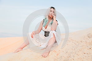 Young beautiful Caucasian woman in white dress and brown leather waistcoat posing in desert landscape with sand.