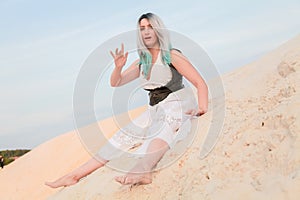 Young beautiful Caucasian woman in white dress and brown leather waistcoat posing in desert landscape with sand.