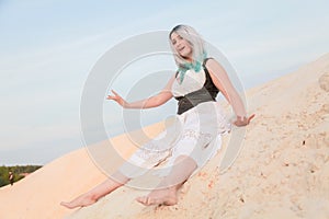 Young beautiful Caucasian woman in white dress and brown leather waistcoat posing in desert landscape with sand.