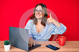 Young beautiful caucasian woman working at the office with laptop smiling happy and positive, thumb up doing excellent and