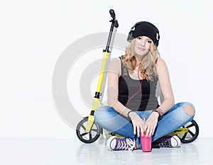 Young beautiful cheerful fashion girl in jeans, sneakers, hat sits on a yellow scooter and listening to music on headphones. Keeps