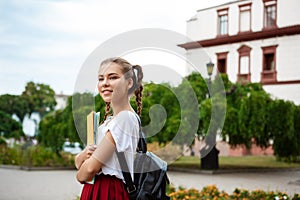 Young beautiful cheerful female student smiling, holding folders outdoors, park background.