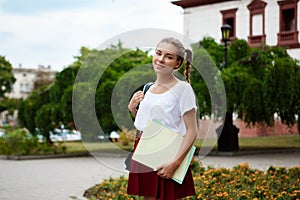 Young beautiful cheerful female student smiling, holding folders outdoors, park background.