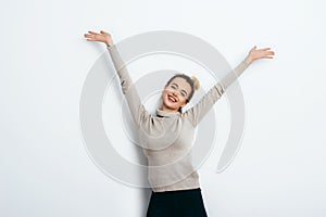 Young beautiful cheerful woman with hair bun wearing in jeans and sweater dancing and posing over white wall. Good mood. Hands up!