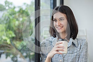 Young beautiful cheerful woman holding paper cup coffee smiling looking out window office. asian business woman relax time