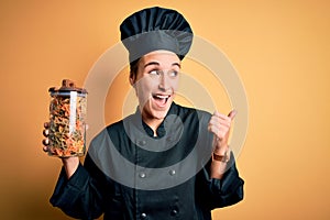 Young beautiful chef woman wearing cooker uniform and hat holding bottle with italian pasta pointing and showing with thumb up to