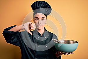 Young beautiful chef woman wearing cooker uniform and hat holding bowl and whisk with angry face, negative sign showing dislike
