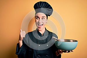 Young beautiful chef woman wearing cooker uniform and hat holding bowl and whisk pointing and showing with thumb up to the side