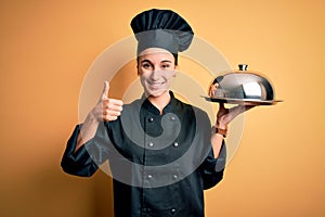 Young beautiful chef woman wearing cooker uniform and hat holding tray with dome happy with big smile doing ok sign, thumb up with