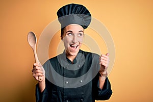 Young beautiful chef woman wearing cooker uniform and hat holding wooden spoon pointing and showing with thumb up to the side with