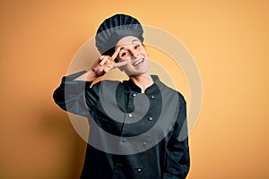 Young beautiful chef woman wearing cooker uniform and hat standing over yellow background Doing peace symbol with fingers over