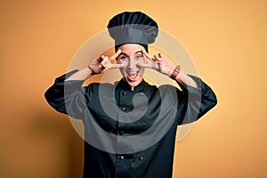 Young beautiful chef woman wearing cooker uniform and hat standing over yellow background Doing peace symbol with fingers over