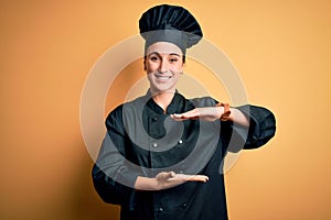 Young beautiful chef woman wearing cooker uniform and hat standing over yellow background gesturing with hands showing big and