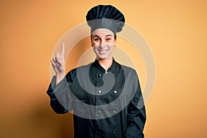 Young beautiful chef woman wearing cooker uniform and hat standing over yellow background showing and pointing up with finger