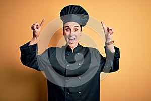 Young beautiful chef woman wearing cooker uniform and hat standing over yellow background smiling amazed and surprised and