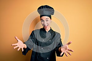 Young beautiful chef woman wearing cooker uniform and hat standing over yellow background smiling cheerful offering hands giving