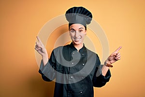 Young beautiful chef woman wearing cooker uniform and hat standing over yellow background smiling confident pointing with fingers