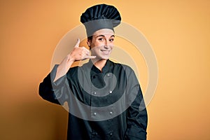 Young beautiful chef woman wearing cooker uniform and hat standing over yellow background smiling doing phone gesture with hand