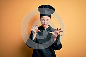 Young beautiful chef woman wearing cooker uniform and hat standing over yellow background smiling funny doing claw gesture as cat,