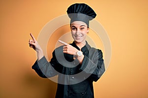 Young beautiful chef woman wearing cooker uniform and hat standing over yellow background smiling and looking at the camera