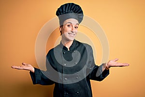 Young beautiful chef woman wearing cooker uniform and hat standing over yellow background smiling showing both hands open palms,