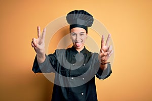 Young beautiful chef woman wearing cooker uniform and hat standing over yellow background smiling with tongue out showing fingers