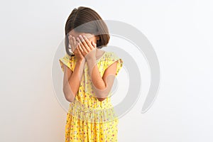 Young beautiful child girl wearing yellow floral dress standing over isolated white background with sad expression covering face