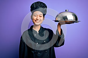 Young beautiful chinese chef woman wearing cooker uniform and hat holding tray with dome with a happy face standing and smiling