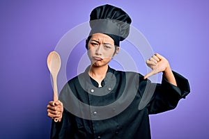 Young beautiful chinese chef woman wearing cooker uniform and hat holding wooden spoon with angry face, negative sign showing