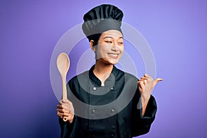 Young beautiful chinese chef woman wearing cooker uniform and hat holding wooden spoon pointing and showing with thumb up to the