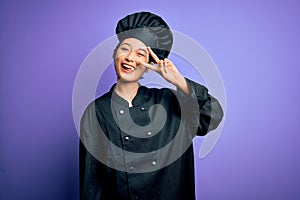 Young beautiful chinese chef woman wearing cooker uniform and hat over purple background Doing peace symbol with fingers over