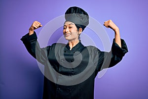 Young beautiful chinese chef woman wearing cooker uniform and hat over purple background showing arms muscles smiling proud