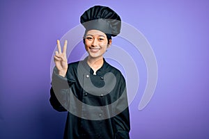 Young beautiful chinese chef woman wearing cooker uniform and hat over purple background smiling looking to the camera showing