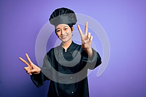 Young beautiful chinese chef woman wearing cooker uniform and hat over purple background smiling with tongue out showing fingers