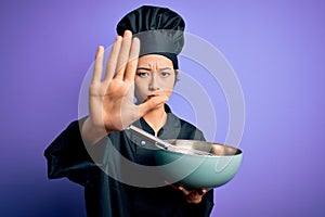 Young beautiful chinese chef woman wearing cooker uniform and hat using bowl and whisk with open hand doing stop sign with serious