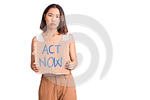 Young beautiful chinese girl holding act now banner thinking attitude and sober expression looking self confident