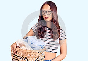 Young beautiful chinese girl holding laundry basket thinking attitude and sober expression looking self confident