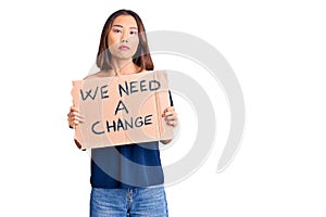Young beautiful chinese girl holding we need a change banner thinking attitude and sober expression looking self confident